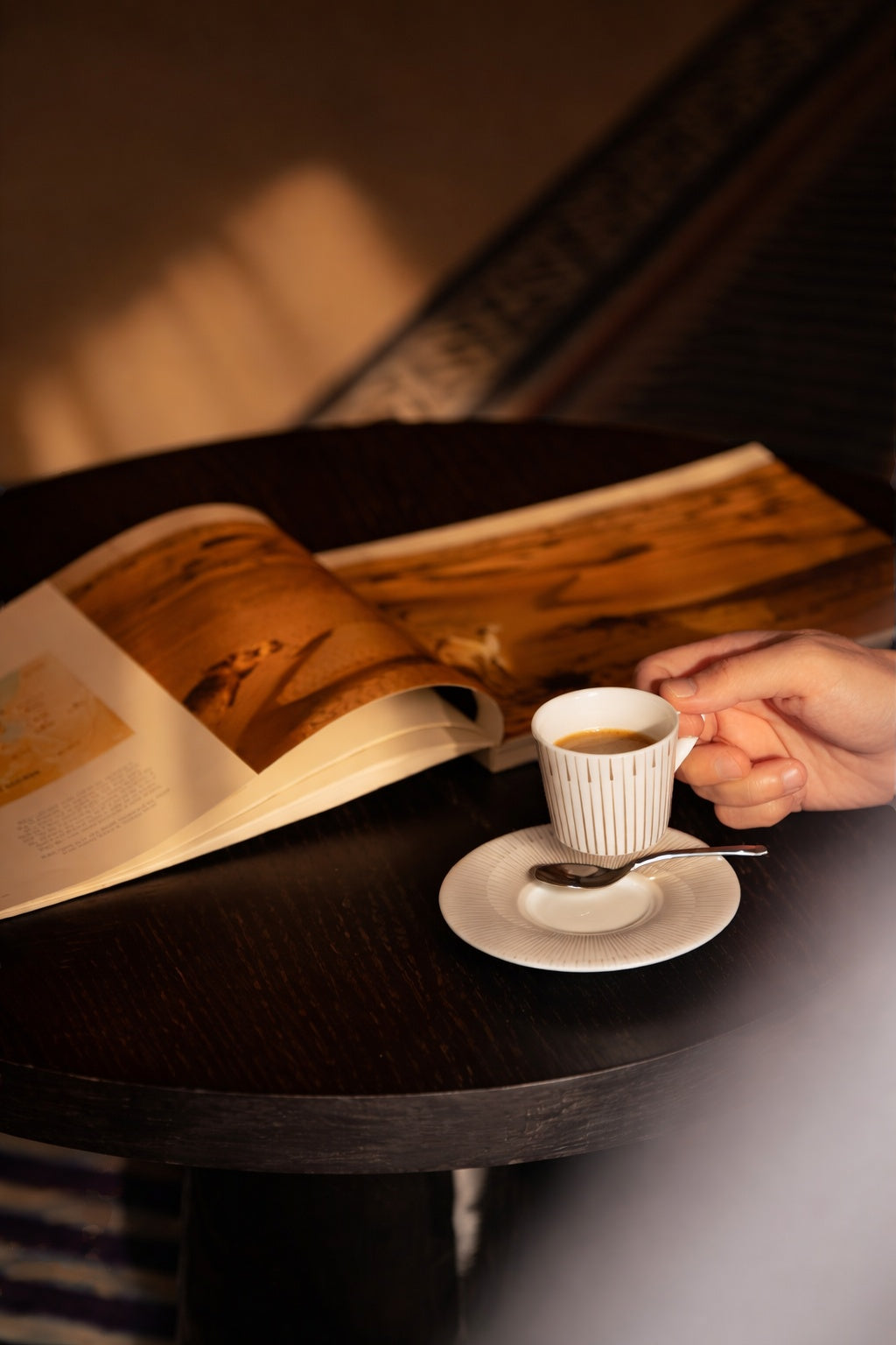 Warm sunlight on a black round table with an open book, espresso cup and saucer held in hand.